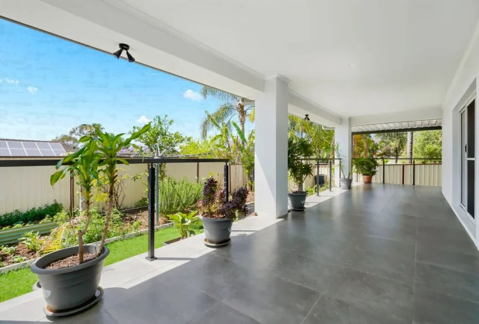 A spacious, covered patio with tiled flooring and potted plants overlooks a well-maintained garden and is enclosed by a white fence, perfect for those seeking supported independent living. SIL Vacancy Blacktown is now available.