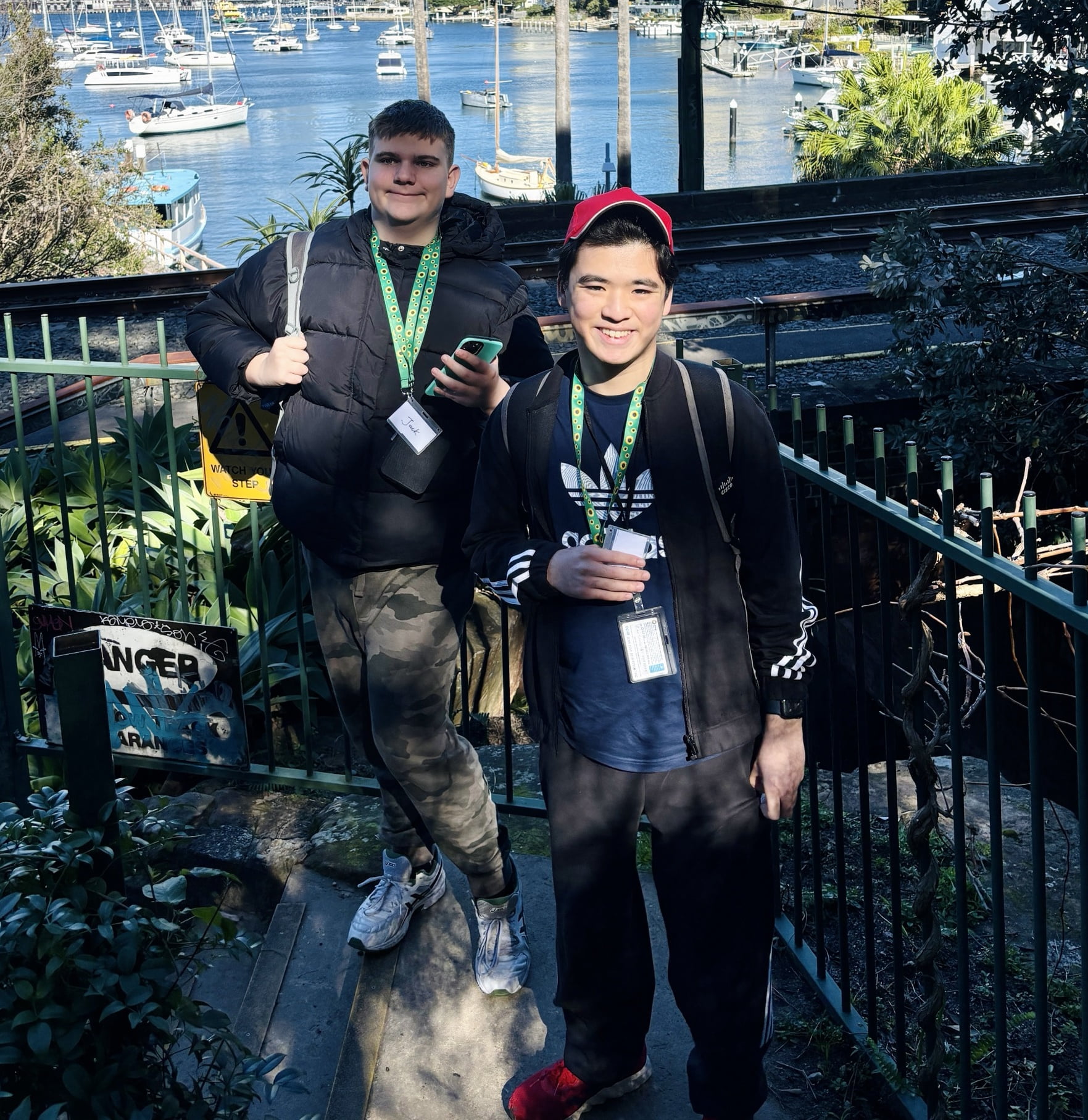 Two people with lanyards and name badges posed on outdoor steps near a marina with moored boats in the background.