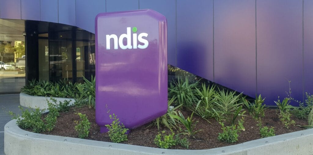 Purple NDIS sign in front of a building with plants at the base, set against a curved, dark blue wall.