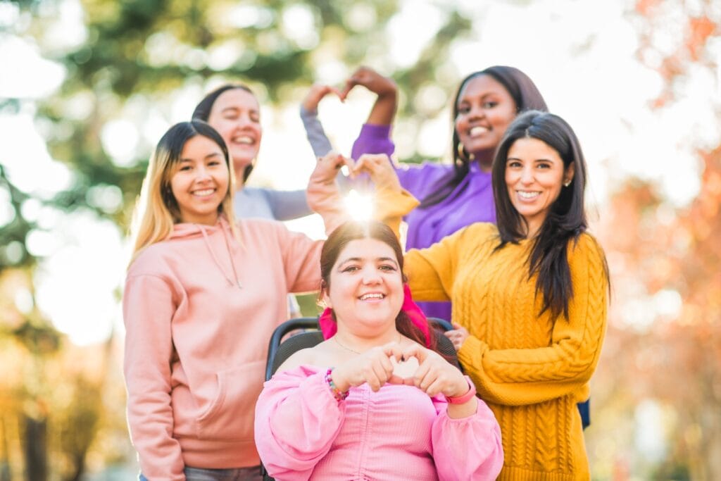 Five women, one in a wheelchair, pose outdoors forming heart shapes with their hands. Their smiles capture the spirit of NDIS social participation, celebrating friendship and inclusion amid trees and sunlight.