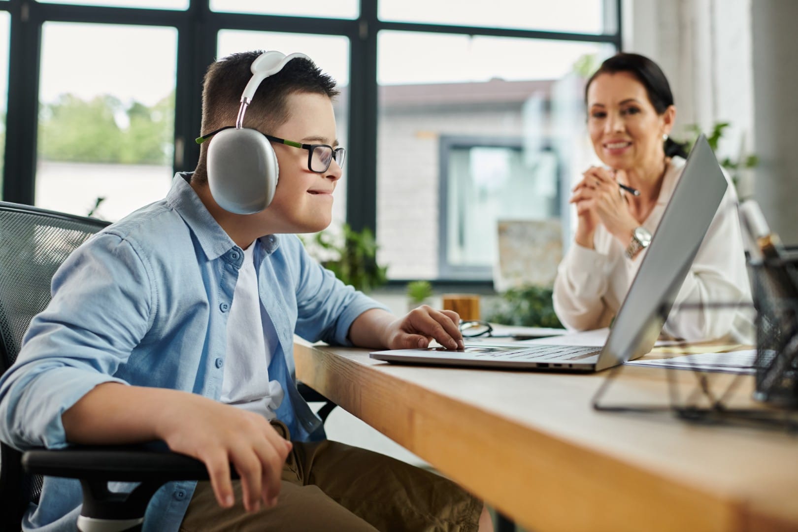 A boy wearing headphones uses a laptop at a desk while a woman sits nearby, watching him and smiling.